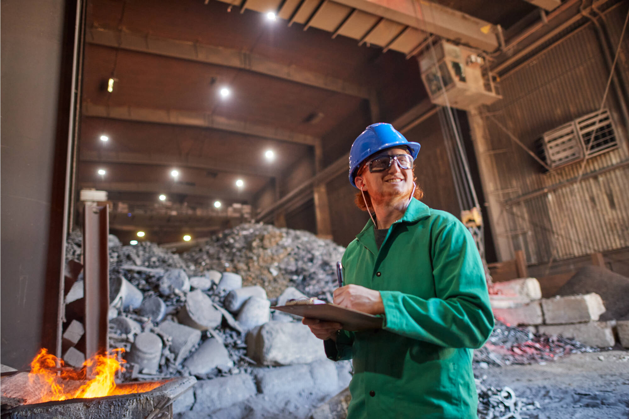 GVSU Student in front of a pile of concrete rubble, wearing green coveralls and a blue hard hat, smiling off into the distance while writing on a clipboard.